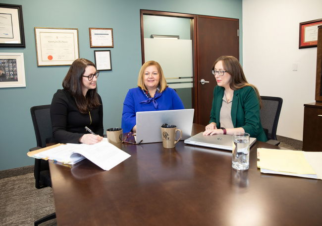 Three women in office reviewing resumes on laptop.